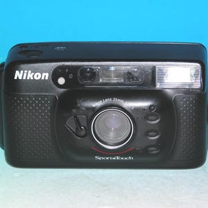 Pode incluir: Câmera de filme Nikon SportTouch 35mm preta. A câmera possui flash embutido, lente e o logotipo Nikon em branco. Possui uma empunhadura texturizada e um detalhe vermelho abaixo da lente.