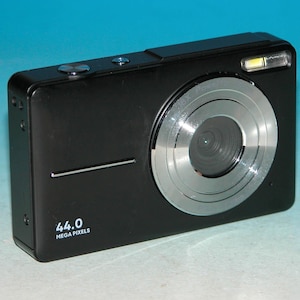 May include: A black digital camera featuring a silver lens barrel, a built-in flash, and a top-mounted shutter button. The front of the camera body displays the text 44.0 MEGA PIXELS in white lettering on the lower left side.