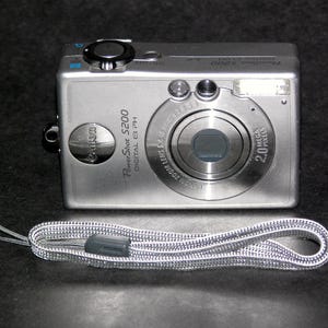 May include: Silver Canon PowerShot S200 digital camera with a matching silver wrist strap. The camera has a lens with a 2.0-megapixel resolution and the text "PowerShot S200 DIGITAL ELPH". The wrist strap is gray and silver.