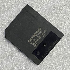 Puede incluir: Una tarjeta SmartMedia negra Olympus M-8P D3V20. La tarjeta tiene el logotipo de Olympus y otro texto impreso, incluyendo "KOREA" y "109Y". El logotipo de SmartMedia también es visible.
