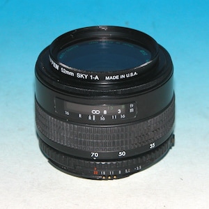 May include: A black camera lens with a blue filter. The lens has a textured grip and markings for focus and aperture. Text on the lens reads "FEN 52mm SKY 1-A MADE IN U.S.A.". The lens has measurements in feet and meters.