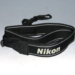 May include: Black camera strap with white stripes and the word "Nikon" printed in white.