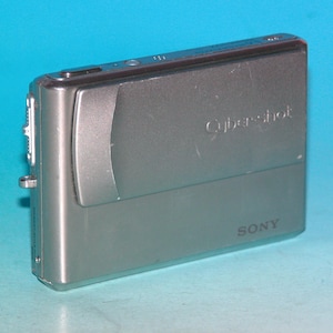 Op de afbeelding: Een zilveren Sony Cybershot digitale camera. De camera heeft een rechthoekige vorm met afgeronde hoeken. Het woord "Cybershot" staat op de voorkant, en het woord "SONY" staat eronder. Op een blauwe achtergrond.