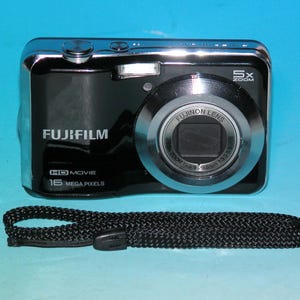 May include: Black Fujifilm digital camera with a silver trim and a black wrist strap. The camera has a 5x zoom and 16 megapixels. The lens is labelled Fujinon Lens.