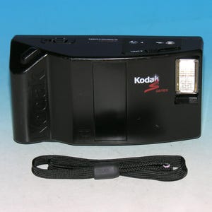 Puede incluir: Cámara Kodak serie S negra con flash incorporado y correa de muñeca negra. La cámara tiene el logotipo de Kodak y la palabra "series" en rojo. La cámara está sobre un fondo azul claro.