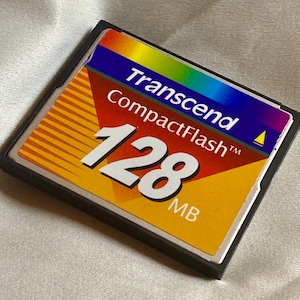 May include: A Transcend CompactFlash memory card, 128 MB capacity. The card has a black frame and a colourful label. The label features the brand name and capacity information, with a gradient of colours including yellow, orange, and blue.