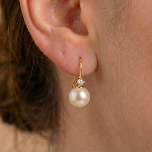 Puede incluir: Un pendiente dorado con una perla blanca y una pequeña gema transparente. El pendiente cuelga de la oreja de una persona. La perla es redonda y tiene una superficie lisa.