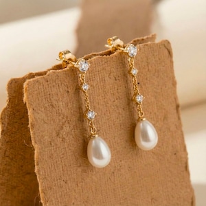 Può includere: Un paio di orecchini pendenti color oro con una catena di piccole pietre chiare e scintillanti. Ogni orecchino ha una perla bianca a forma di goccia nella parte inferiore. Gli orecchini sono esposti su una superficie marrone strutturata.