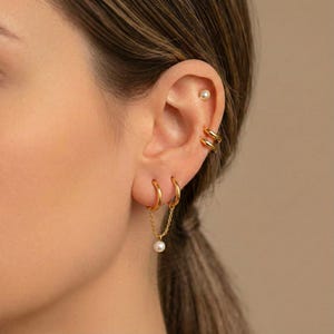 Könnte beinhalten: Nahaufnahme eines Ohrs mit goldenen Ohrringen. Das Ohr zeigt einen kleinen Perlenstecker, zwei gestapelte Ohrklemmen und zwei Creolen, eine davon mit einer Kette und einem hängenden Perlenanhänger. Der Schmuck hat ein glänzendes Finish.