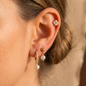 May include: An ear featuring three pieces of jewellery. A gold flower stud earring is on the upper ear. A gold hoop earring with small clear stones and a pearl drop earring are on the lower ear. A gold ball stud earring with a pearl drop is also visible.