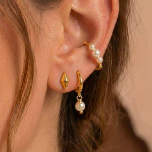 May include: A close-up of a person's ear adorned with gold earrings. The set includes a diamond-shaped stud, a small hoop with a dangling pearl, and a gold ear cuff with three pearls. The jewelry has a polished, reflective finish.