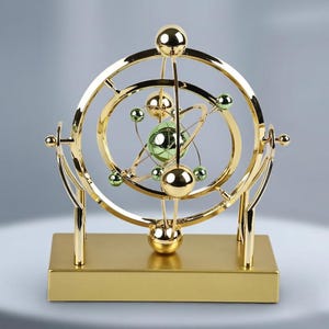 Pode incluir: Uma escultura cinética dourada com um design esférico, apresentando esferas em órbita e uma esfera verde central. A escultura é montada em uma base retangular e tem um acabamento polido, criando uma exibição decorativa e dinâmica.