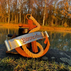 Könnte beinhalten: Ein orangefarbener und brauner Anker mit einem Seil darum und einem Banner mit der Aufschrift "Binnenband" in einer dekorativen Schriftart. Der Anker befindet sich vor dem Hintergrund eines ruhigen Gewässers und Bäumen.