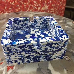 May include: Square ashtray with a blue and white speckled pattern. The ashtray has a rectangular shape with a recessed area for holding cigarettes. The background is a red and white speckled surface.
