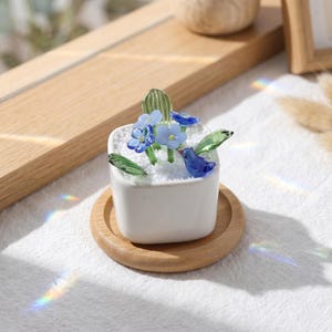 May include: A small, white square ceramic pot with a miniature glass garden. The garden features blue glass flowers, green leaves, and a blue glass bird. The pot sits on a round wooden coaster.