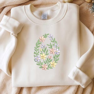Könnte beinhalten: Cremefarbenes Sweatshirt mit einem gestickten floralen Eierdesign. Das Ei ist mit pastellfarbenen Blumen und grünen Blättern gefüllt. Das Sweatshirt hat einen Rundhalsausschnitt und lange Ärmel mit gerippten Bündchen.