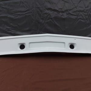 Peut inclure: Un pare-chocs de voiture blanc avec deux trous circulaires et une encoche rectangulaire. Le pare-chocs a un design incurvé et est probablement fait d'un matériau durable. Le fond est un tissu gris foncé.