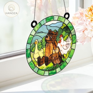 May include: A stained glass window featuring a bear, duck, owl, and chicken in a forest scene. The artwork is framed in green and hangs from a black chain. The HANOZA logo is in the upper left corner.