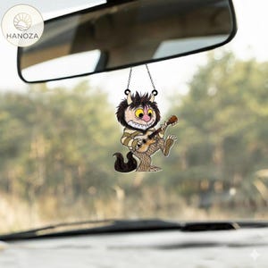 Op de afbeelding: Een auto-accessoire met een stripfiguur die gitaar speelt. De charme hangt aan een achteruitkijkspiegel. Het personage heeft bruine vacht, een gestreept shirt en visnetkousen. De achtergrond is een wazig uitzicht op bomen.
