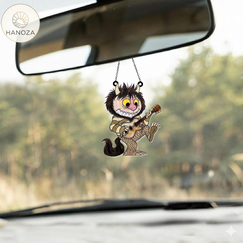 Op de afbeelding: Een auto-accessoire met een stripfiguur die gitaar speelt. De charme hangt aan een achteruitkijkspiegel. Het personage heeft bruine vacht, een gestreept shirt en visnetkousen. De achtergrond is een wazig uitzicht op bomen.
