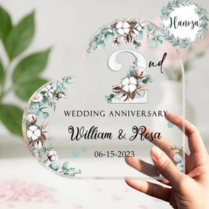 Op de afbeelding: Een heldere, hartvormige trouwjubileumplaquette van acryl. De plaquette heeft bloemaccenten, de woorden "2nd Wedding Anniversary", de namen "William & Rosa" en de datum "06-15-2023".