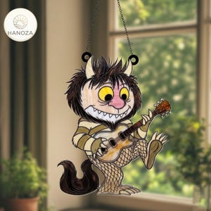 Op de afbeelding: Gebrandschilderde zonnevanger met een cartoonmonster dat gitaar speelt. Het monster heeft bruin haar, gele ogen en een roze neus. Het draagt een gestreept shirt en heeft een lange, donkere staart. De zonnevanger hangt aan een ketting en heeft de merknaam "HANOZA".