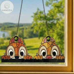 May include: Stained glass window hangings featuring Chip and Dale, the Disney chipmunks. The artwork shows the chipmunks peeking over a black ledge, with brown, tan, and white details. Each piece is suspended by a chain.