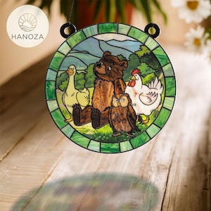 May include: Round stained glass panel featuring a bear, owl, duck, and chicken in a pastoral scene. The panel has a green border and is suspended by a black chain. The HANOZA logo is visible in the upper left corner.