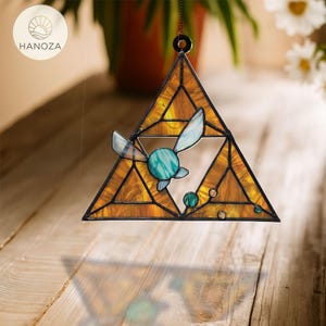 May include: A stained glass triangle suncatcher with a geometric design in shades of amber and gold. It features a teal and blue fairy-like figure in the center and small teal orbs. The suncatcher is suspended by a chain.