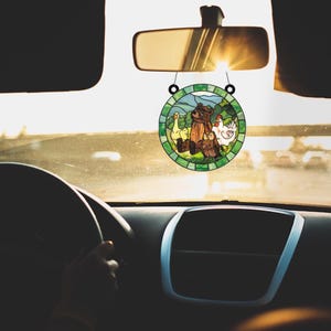 May include: A round, stained-glass car charm featuring a bear, two owls, a duck, and a chicken in a pastoral scene. The charm is suspended from a car's rearview mirror, with the sun shining through the windshield.