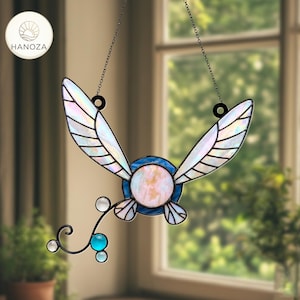 Peut inclure: Suncatcher en vitrail avec un motif de fée. Il présente des ailes irisées, un globe central rose et blanc, et un bord bleu. Une chaîne décorative et un petit fil incurvé avec des perles bleues et transparentes complètent le design.