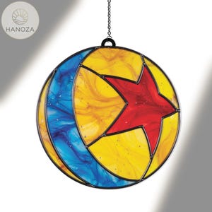 Puede incluir: Un atrapasoles redondo de vidriera con un marco de metal negro. El dise&ntilde;o presenta una estrella roja en el centro, rodeada de secciones amarillas y azules. Una cadena est&aacute; unida para colgarlo. Un art&iacute;culo decorativo.