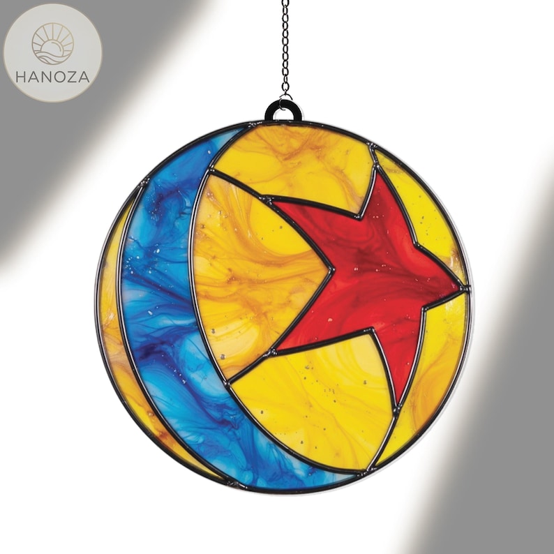 Puede incluir: Un atrapasoles redondo de vidriera con un marco de metal negro. El dise&ntilde;o presenta una estrella roja en el centro, rodeada de secciones amarillas y azules. Una cadena est&aacute; unida para colgarlo. Un art&iacute;culo decorativo.