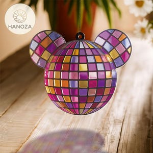 Op de afbeelding: Een decoratief muisvormig ornament, dat lijkt op een discobal. Het ornament bestaat uit kleine, vierkante tegels in tinten paars, roze, oranje en goud. Het hangt aan een ketting. De oren zijn cirkelvormig en ook betegeld.