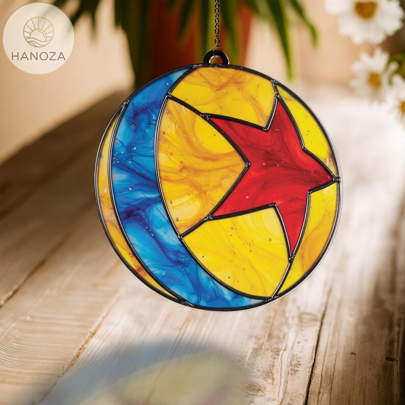 Puede incluir: Un atrapasoles redondo de vidriera con un marco de metal negro. El dise&ntilde;o presenta una estrella roja, una media luna azul y un fondo amarillo. El atrapasoles est&aacute; colgado de una cadena. La marca "HANOZA" es visible.