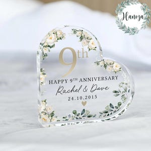 Puede incluir: Recuerdo de aniversario en forma de corazón de acrílico transparente con detalles florales. El diseño presenta el texto "9th", "Happy 9th Anniversary", los nombres "Rachel & Dave" y la fecha 24.10.2015. Decorado con flores blancas y hojas verdes.