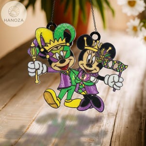 May include: Two colorful suncatchers featuring cartoon characters in Mardi Gras attire. One character wears a jester hat and holds a scepter, while the other holds a mask. The suncatchers are in shades of green, purple, yellow, and black.