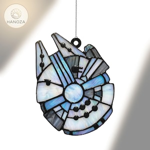 May include: A stained glass suncatcher shaped like the Millennium Falcon. It has a black border and is filled with white, blue, and grey glass. It is suspended by a small chain and ring.