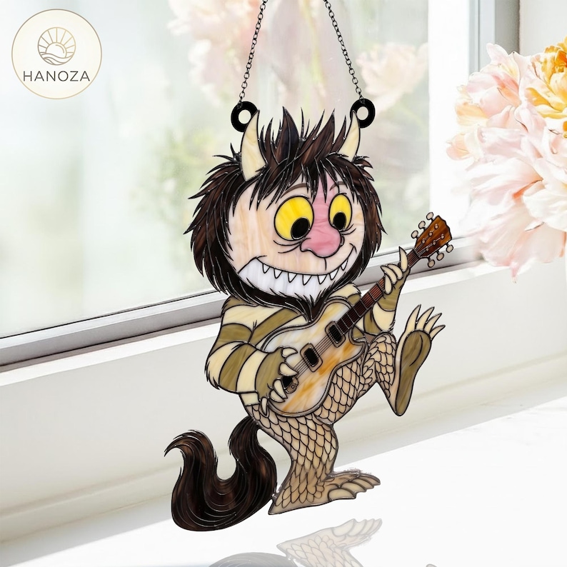 Op de afbeelding: Een gebrandschilderde raamdecoratie met een cartoonmonster dat gitaar speelt. Het monster heeft gele ogen, een roze neus en een gestreept shirt. De gitaar is bruin en de staart van het monster is zwart. Het hangt aan een zwarte ketting.