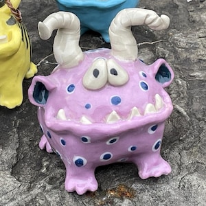 May include: Three whimsical ceramic monster figurines in yellow, blue, and pink. Each has large eyes, horns, and a toothy grin. The monsters are posed on a gray stone surface, surrounded by greenery.