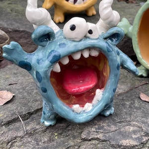 Könnte beinhalten: Drei skurrile Keramik-Monsterfiguren in Blau, Gelb und Grün. Jedes hat einen weit geöffneten Mund mit roter Zunge, weißen Zähnen und Hörnern. Die Monster werden auf einem grauen Felsen mit grünem Laub im Hintergrund gezeigt.