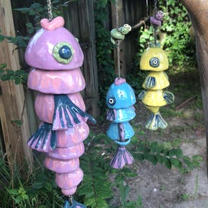 May include: Three ceramic fish wind chimes in shades of pink, blue, and yellow hang from a chain. The fish have layered, rounded bodies and detailed fins. The background includes a wooden fence, tree, and greenery.