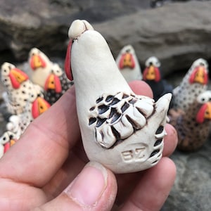 May include: A white ceramic chicken figurine with black and brown detailing. The chicken is standing upright with its head tilted back. The figurine is hand-painted and has a unique, rustic style.