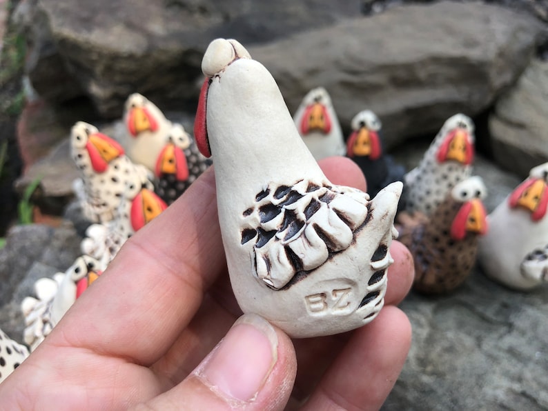 May include: A white ceramic chicken figurine with black and brown detailing. The chicken is standing upright with its head tilted back. The figurine is hand-painted and has a unique, rustic style.