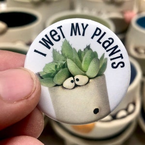 May include: White button with black text that reads "I wet my plants". The button features a cartoon succulent plant with googly eyes in a white pot.
