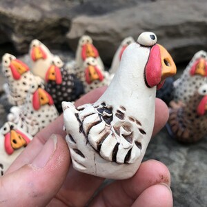 May include: A collection of white ceramic chickens with black and brown accents. The chickens have large, round eyes and red beaks. One chicken is being held in a hand.