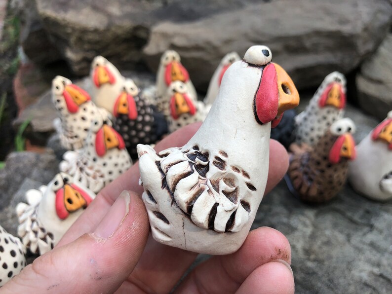 May include: A collection of white ceramic chickens with black and brown accents. The chickens have large, round eyes and red beaks. One chicken is being held in a hand.