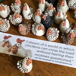 May include: A collection of hand-painted ceramic chickens with black and white polka dots, brown and white, and black and white patterns. The chickens are arranged on a wooden surface with a sign that reads "I dream of a world in which chickens can cross the road without having their motives questioned." Join the movement at www.Beckyzee.com