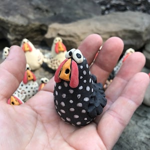 May include: A black and white speckled ceramic chicken figurine with a red beak and yellow nostrils. The chicken is standing on a hand.