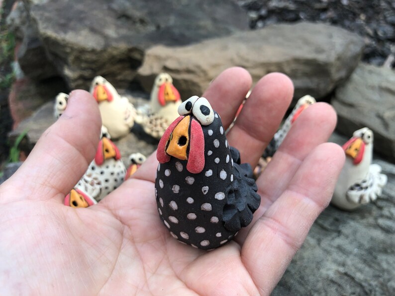 May include: A black and white speckled ceramic chicken figurine with a red beak and yellow nostrils. The chicken is standing on a hand.